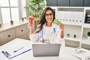 Young hispanic doctor woman holding anatomical female genital organ and birth control pills winking...