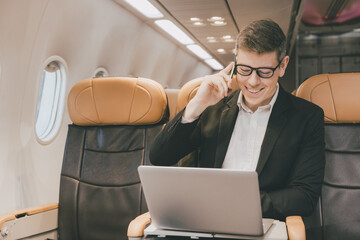 Businessman using satellite internet on laptop and phone calling business contact during air flight...