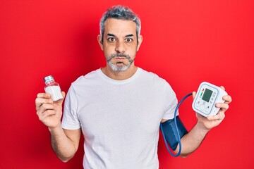 Handsome middle age man with grey hair using blood pressure monitor holding salt puffing cheeks...