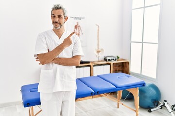 Middle age hispanic therapist man working at pain recovery clinic pointing with hand finger to the side showing advertisement, serious and calm face