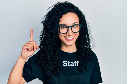 Young hispanic woman with curly hair wearing staff t shirt pointing finger up with successful idea. exited and happy. number one.