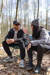 Young man and woman sitting on chairs in forest and having a conversation. Friendship, joy, social life and smiling concept. They are drinking tea. Blur trees background. Vertical photo.