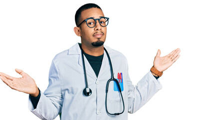 Young african american man wearing doctor uniform and stethoscope clueless and confused expression with arms and hands raised. doubt concept.