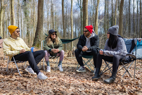 Four friends sitting on camp chairs in forest. They are talking with each others. Friendship, camping, nature and happiness concept. Social and active lifestyle.