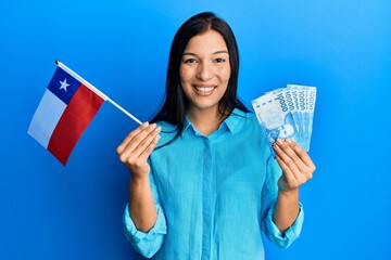 Young latin woman holding chile flag and chilean pesos banknotes smiling with a happy and cool smile on face. showing teeth. - obrazy, fototapety, plakaty