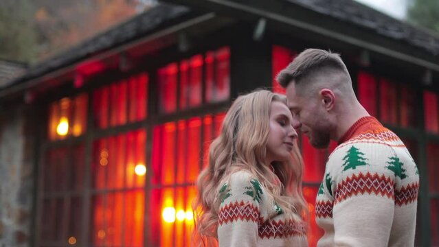 Romantic couple standing hugging near the country house with red lights inside. Young people dating and kissing outdoors.