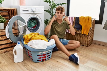 Young caucasian man putting dirty laundry into washing machine covering ears with fingers with annoyed expression for the noise of loud music. deaf concept.