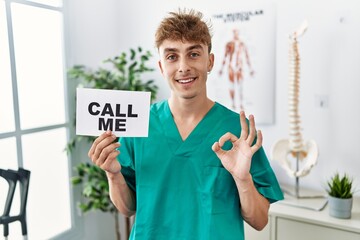 Young caucasian physio man holding call me banner at the clinic doing ok sign with fingers, smiling...