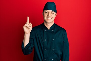 Handsome young man wearing professional cook uniform and hat showing and pointing up with finger number one while smiling confident and happy.