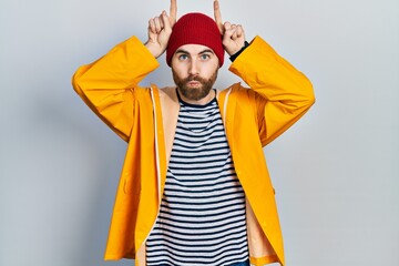Caucasian man with beard wearing yellow raincoat doing funny gesture with finger over head as bull horns