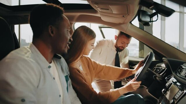 Caucasian car dealer in formal outfit presenting new model of luxury auto to multicultural couple at showroom. Happy customers sitting in car and examining all details of interior. Salesman presenting