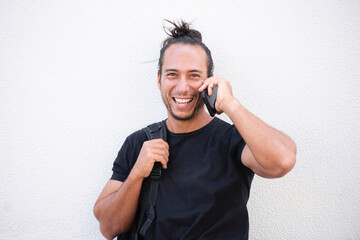 Cheerful man holding backpack and talking on mobile phone outdoors in city