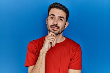 Young hispanic man with beard wearing red t shirt over blue background with hand on chin thinking about question, pensive expression. smiling with thoughtful face. doubt concept.