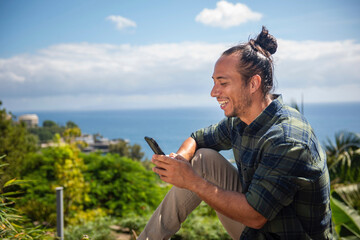 Happy man traveler with pulled up hair bun text messaing using mobile phone overlooking sea