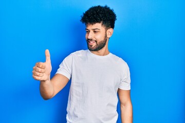 Young arab man with beard wearing casual white t shirt looking proud, smiling doing thumbs up gesture to the side