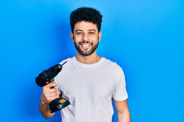 Young arab man with beard using drill looking positive and happy standing and smiling with a...