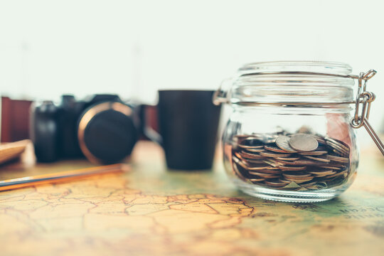 Travel money savings in a glass jar, camera, coffee cup, straw hat and pen on world map. Travel budget concept.