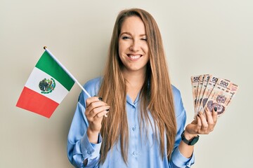 Young blonde woman holding mexico flag and mexican pesos banknotes winking looking at the camera...