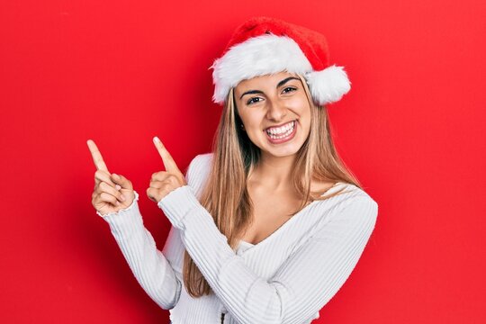 Beautiful hispanic woman wearing christmas hat smiling and looking at the camera pointing with two hands and fingers to the side.