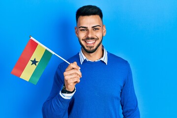 Young hispanic man with beard holding ghana flag looking positive and happy standing and smiling with a confident smile showing teeth