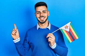 Young hispanic man with beard holding zimbabwe flag smiling happy pointing with hand and finger to the side