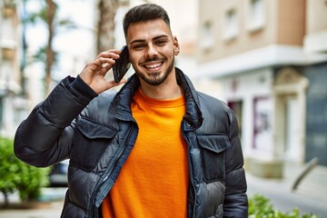 Handsome hispanic man with beard smiling happy and confident at the city wearing winter coat speaking on the phone