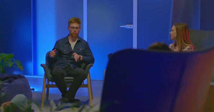 Red-haired guy sits in a chair and talks with colleagues, meeting partners. Guy with glasses talks about a new business project, a modern coworking office. 4k, ProRes