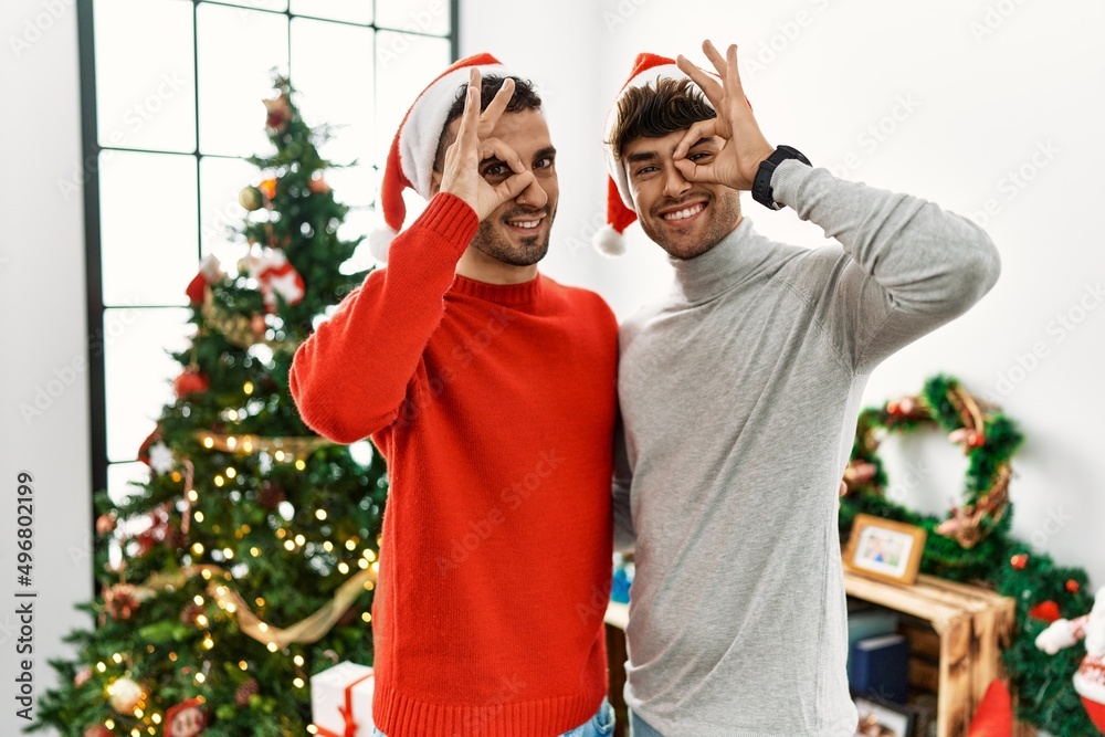 Poster Young gay couple standing by christmas tree wearing hat doing ok gesture with hand smiling, eye looking through fingers with happy face.