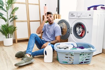 Young hispanic man putting dirty laundry into washing machine doing ok gesture shocked with surprised face, eye looking through fingers. unbelieving expression.