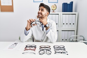 Young optician man holding optometry glasses looking away to side with smile on face, natural...