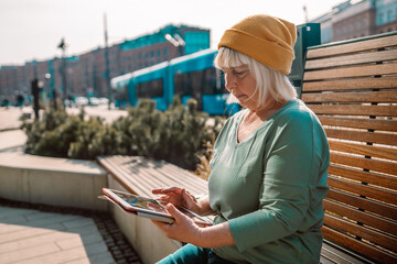 50s 60s Woman using digital tablet PC sitting on a modern wood bench in city walking