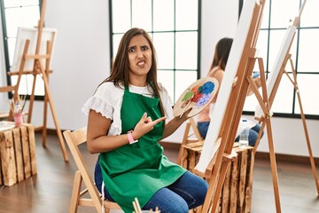 Young hispanic artist women painting on canvas at art studio pointing aside worried and nervous...