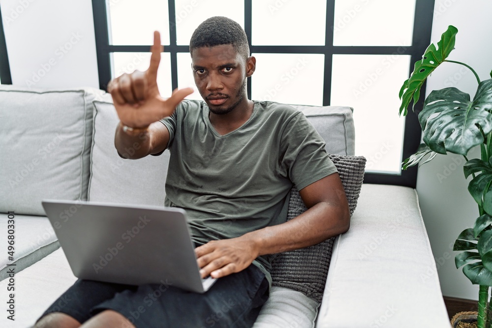 Sticker Young african american man using laptop at home sitting on the sofa pointing with finger up and angry expression, showing no gesture