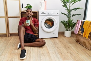 Young african american man using smartphone waiting for washing machine thinking concentrated about...