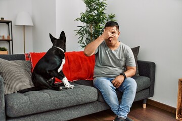 Young latin man and dog sitting on the sofa at home peeking in shock covering face and eyes with hand, looking through fingers with embarrassed expression.