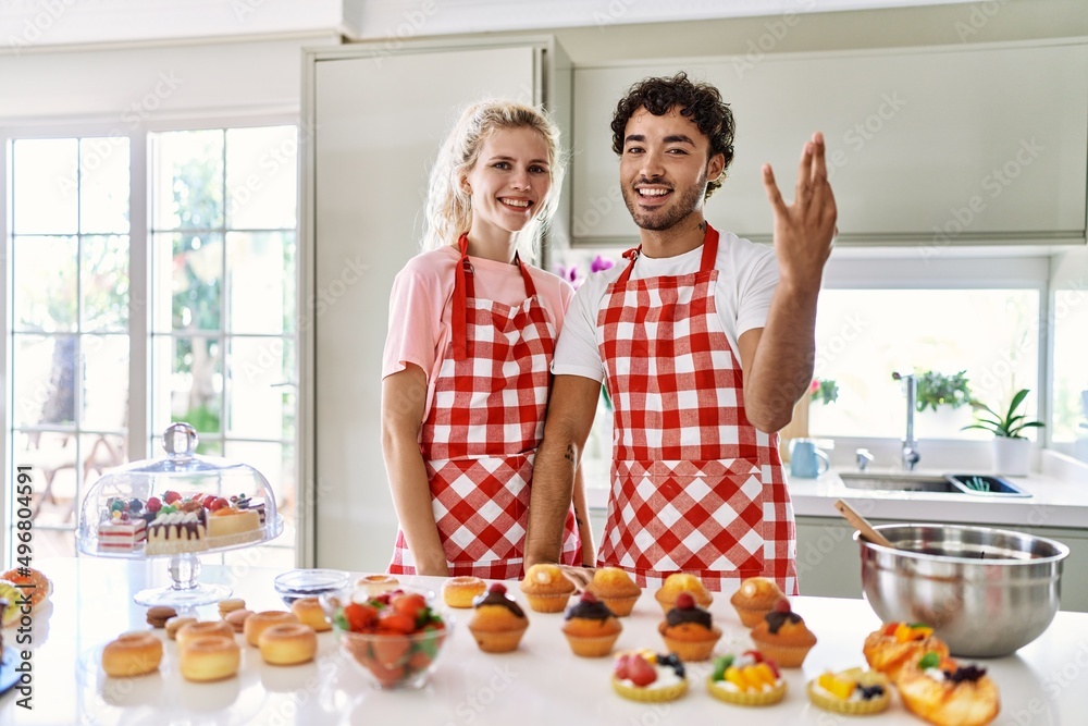 Canvas Prints Couple of wife and husband cooking pastries at the kitchen showing and pointing up with fingers number three while smiling confident and happy.