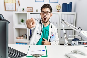 Young man with beard wearing doctor uniform and stethoscope at the clinic pointing with finger surprised ahead, open mouth amazed expression, something on the front