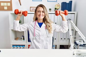 Young beautiful doctor woman holding dumbbells for sport therapy winking looking at the camera with...