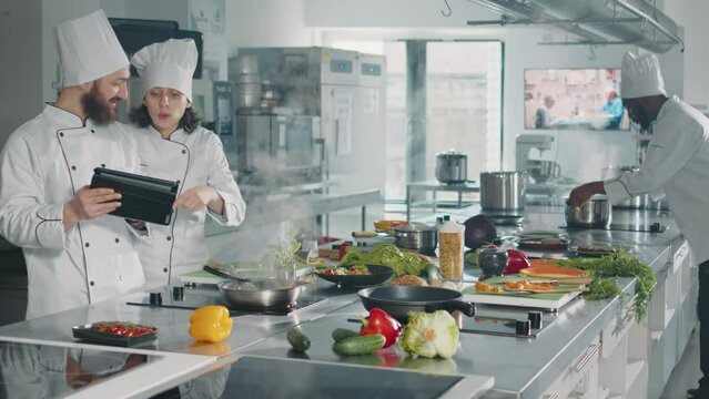 Man and woman analyzing culinary recipe on digital tablet, using internet website to make professional food menu in restaurant kitchen. Team of chefs cooking gastronomic dish with gadget.