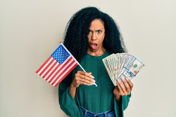 Middle age african american woman holding united states flag and dollars in shock face, looking skeptical and sarcastic, surprised with open mouth
