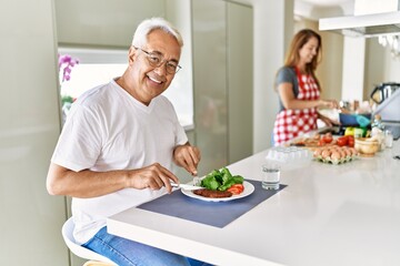 Middle age hispanic man smiling happy eating beef with salad while woman cook at the kitchen.
