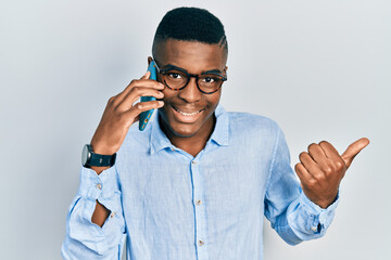 Young african american man having conversation talking on the smartphone pointing thumb up to the side smiling happy with open mouth