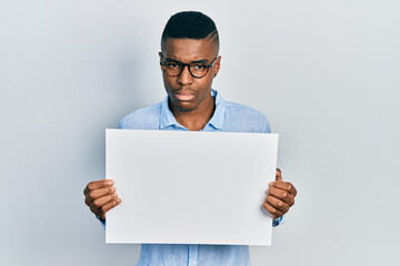 Young african american man wearing glasses holding blank empty banner clueless and confused expression. doubt concept.