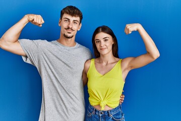 Young hispanic couple standing together over blue background strong person showing arm muscle,...