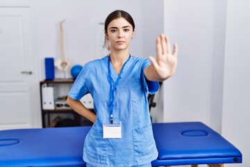Young hispanic woman wearing physiotherapist uniform standing at clinic doing stop sing with palm...