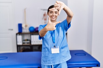 Young hispanic woman wearing physiotherapist uniform standing at clinic smiling making frame with hands and fingers with happy face. creativity and photography concept.