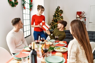 Group of young people smiling happy having christmas dinner. Woman giving gift to man at home.