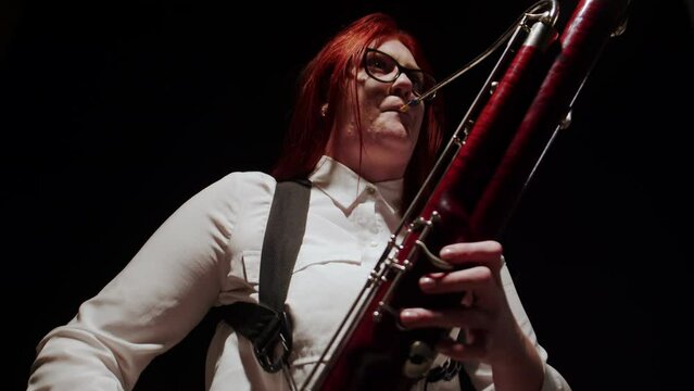 Woman musician plays classical music on bassoon while sitting on chair in studio, dark background, front view. Female musician plays music at bassoon. Concept music