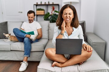 Hispanic middle age couple at home, woman using laptop touching mouth with hand with painful expression because of toothache or dental illness on teeth. dentist