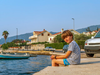 Fototapeta na wymiar A boy in a striped t-shirt and swimming trunks sits on the seashore and looks at the camera.